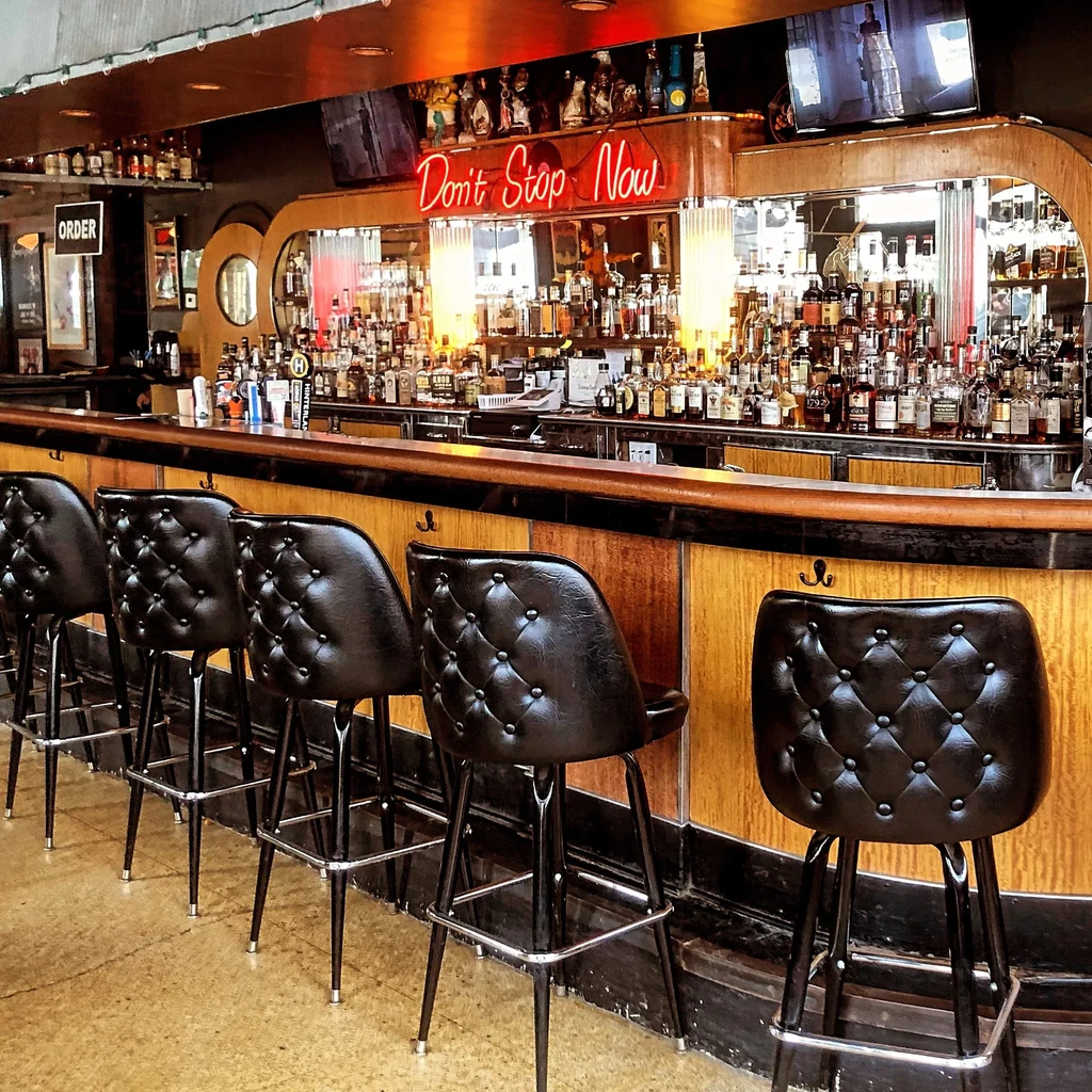 Dimly lit bar, empty stools, rows of bottles, "Don't Stop Now" neon.