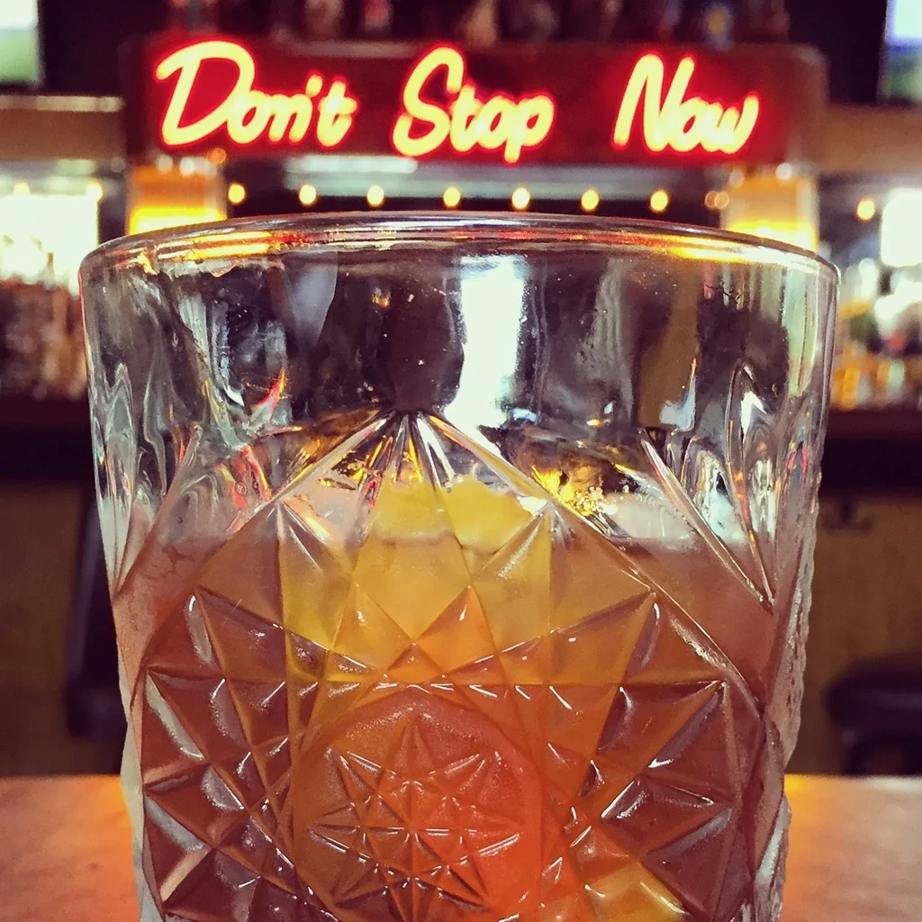 Amber drink in faceted glass, ice, orange peel, 'Don't Stop Now' sign.