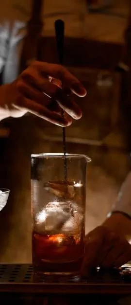 Hands stirring an amber cocktail with ice in a bar mixing glass.