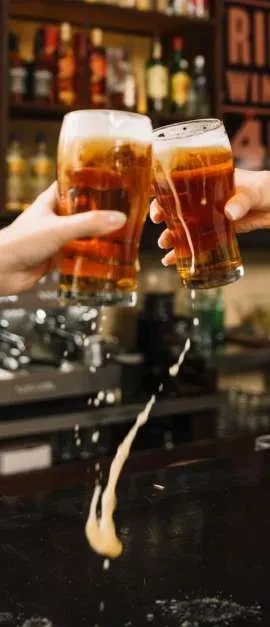 Two hands clink frothy beer pints, spilling amber liquid and foam.