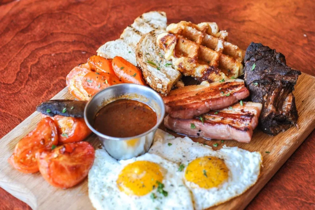 Hearty brunch platter with eggs, bacon, waffles, toast, and savory meat.