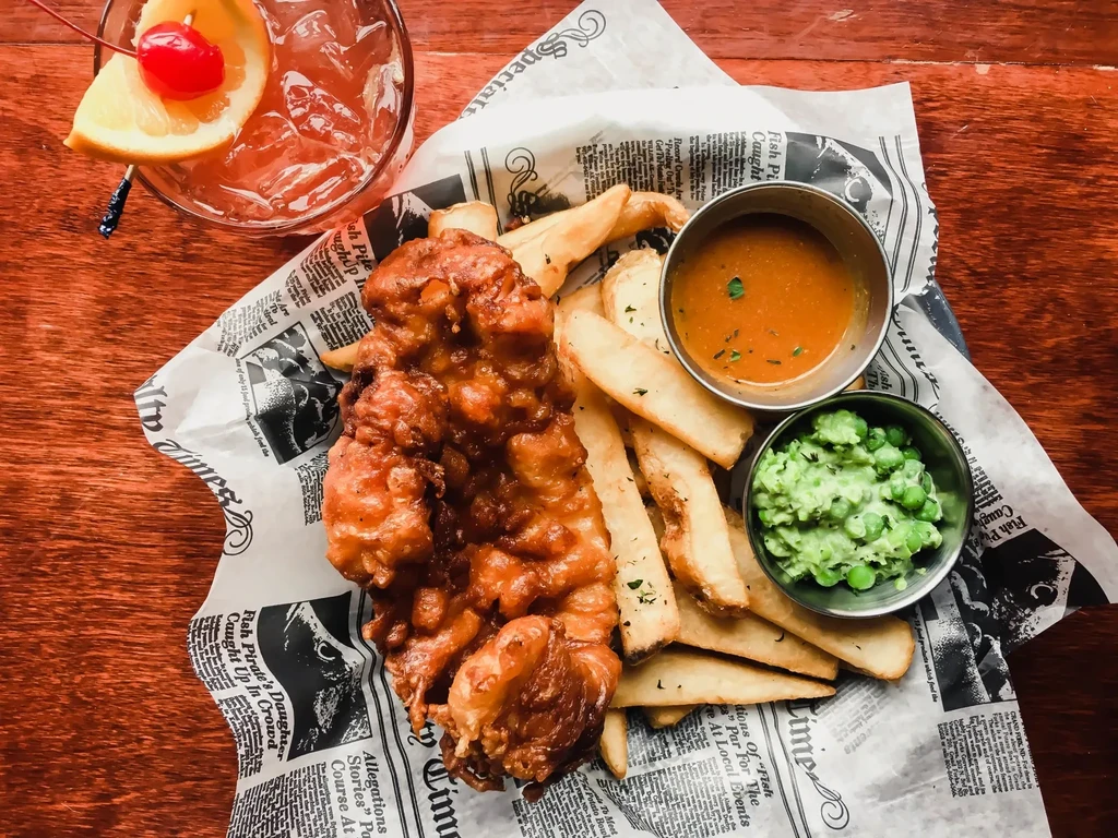 Fish and chips, mushy peas, sauce, and iced drink on wooden table.