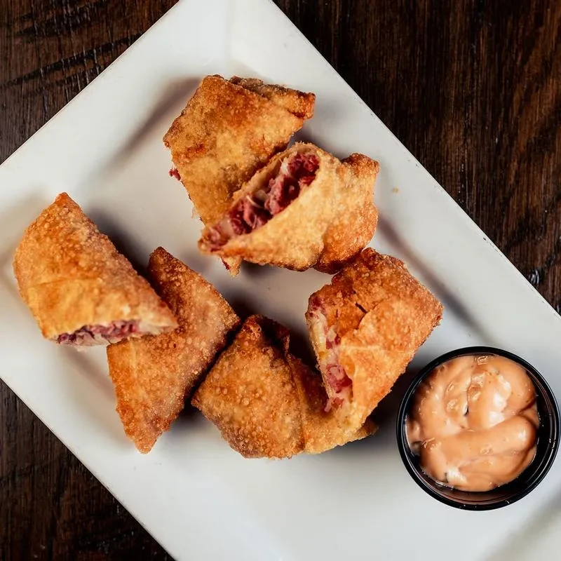 Golden-brown corned beef spring rolls, some cut open, with dipping sauce.