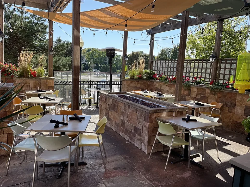 Sunny outdoor dining patio with fire pit, pergola, and colorful chairs.