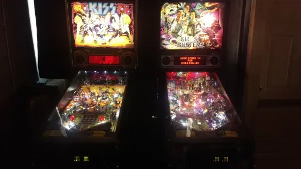 Two illuminated KISS and Ghostbusters pinball machines in a dim room.