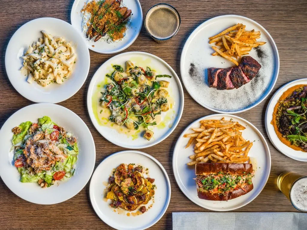 Wooden table flat lay showing nine varied dishes and two beers.