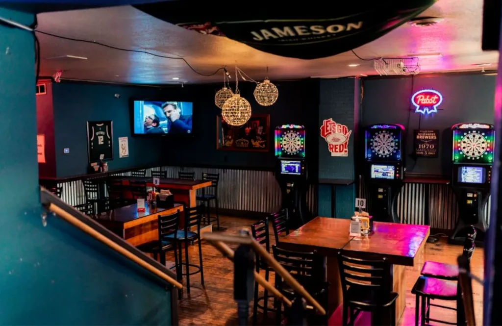 Empty, dimly lit bar with electronic dartboards, neon signs, and TV.