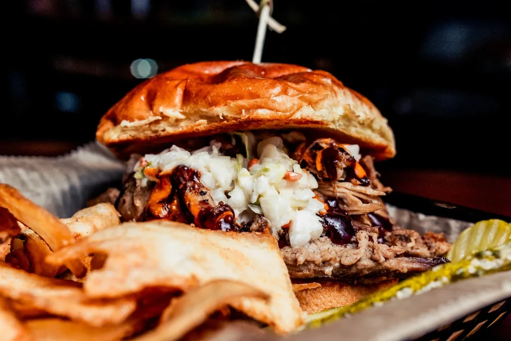 Close-up of a large burger with coleslaw, BBQ sauce, fries, and pickles.
