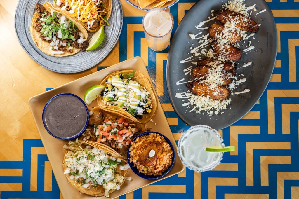 Overhead view of colorful tacos, plantains, and drinks on a patterned table.