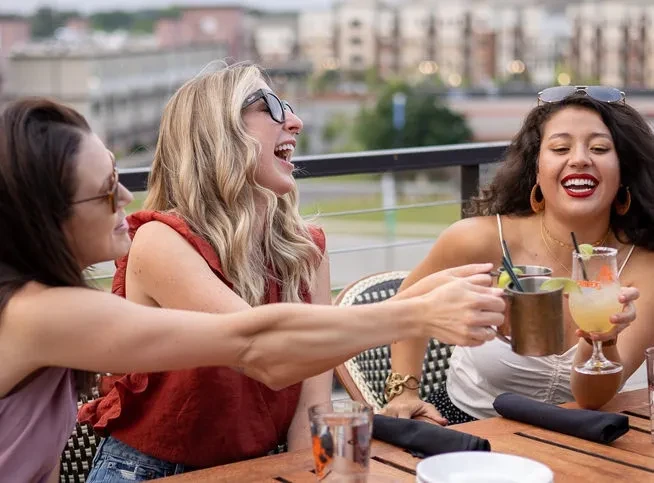 Three joyful women share drinks on an urban rooftop patio.