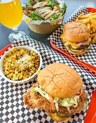 Tabletop spread of casual food: fried chicken, burger, salad, corn, drink.