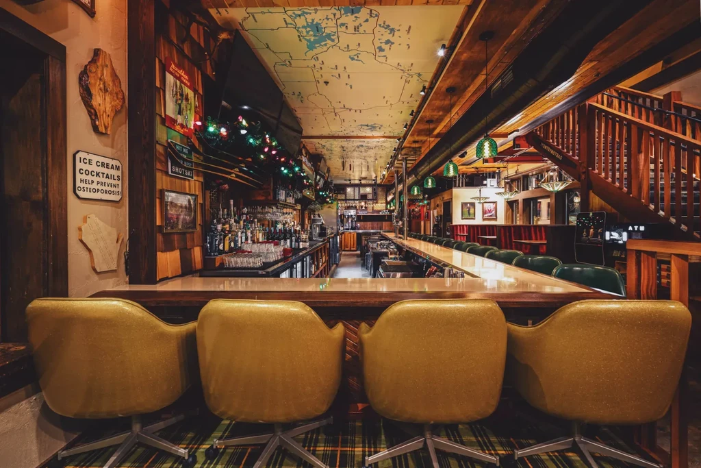 Rustic bar interior: empty yellow stools, stocked bar, map ceiling.