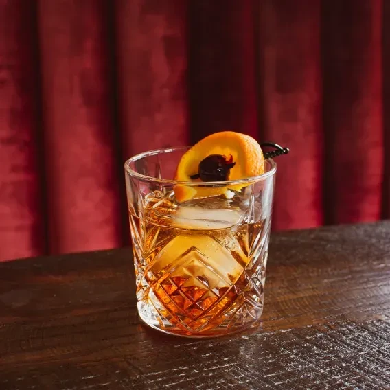 Amber cocktail with spherical ice, orange twist, and cherry on wood.