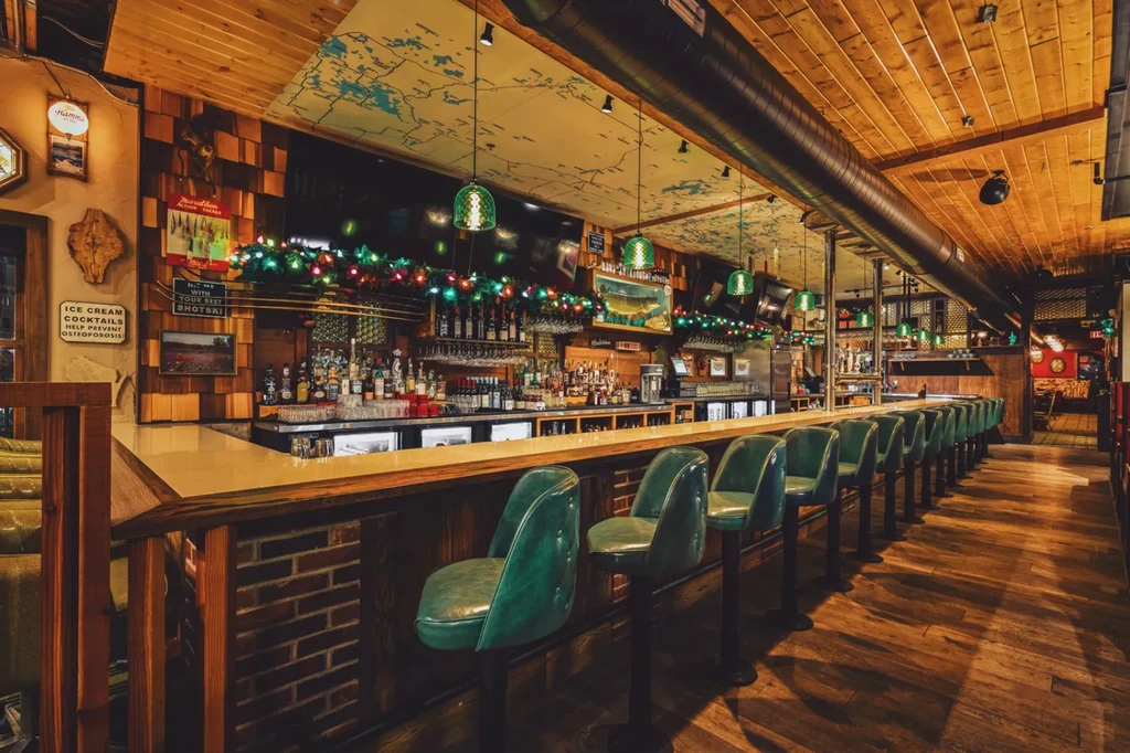 Rustic bar interior with green stools, liquor bottles, and festive lights.