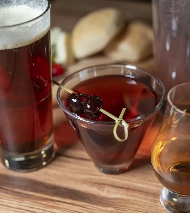 Red cocktail with cherries, beer, and amber spirit on a wooden table.