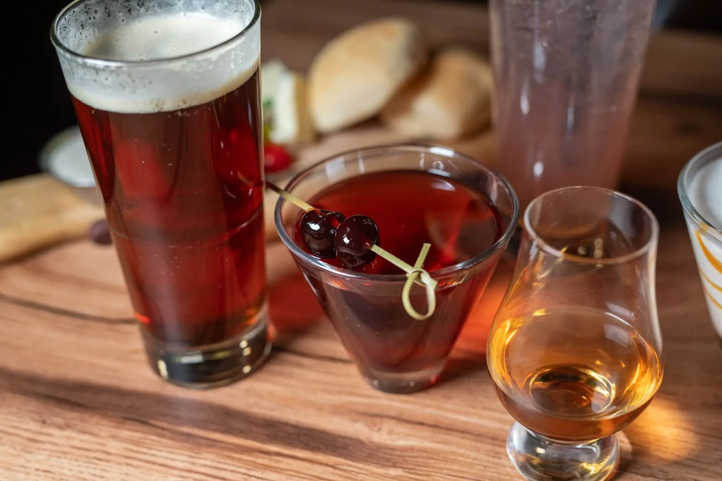 Beer, red cocktail, whisky, and appetizers on warm wooden surface.