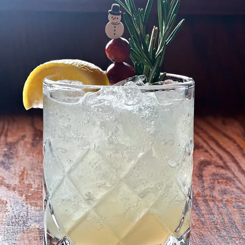 Bubbly drink in diamond glass, garnished with lemon, berries, snowman, rosemary.