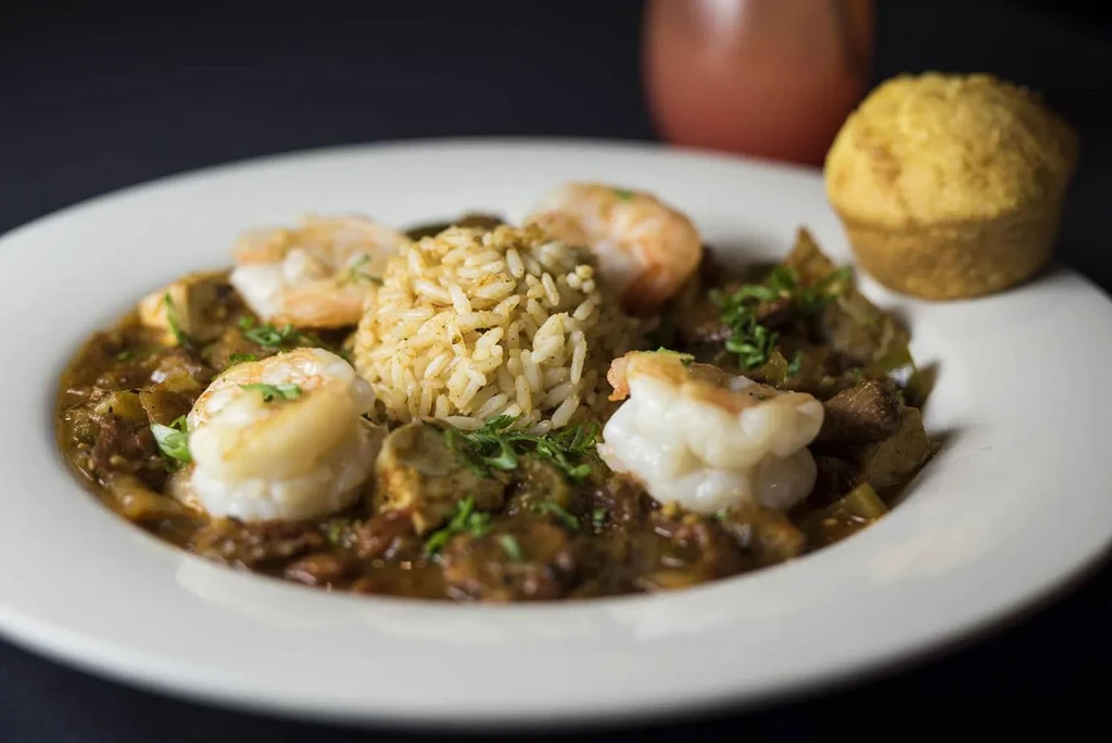 Close-up of a dark stew with rice, shrimp, herbs, and cornbread.