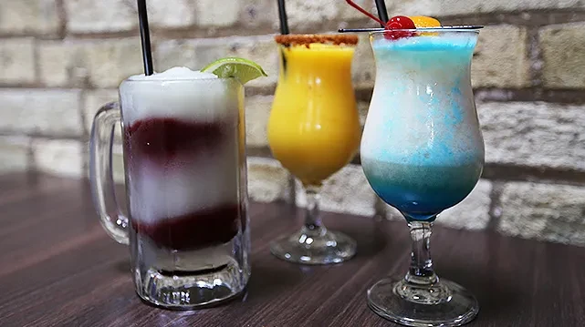 Three colorful frozen drinks with varied layers and garnishes on wood.