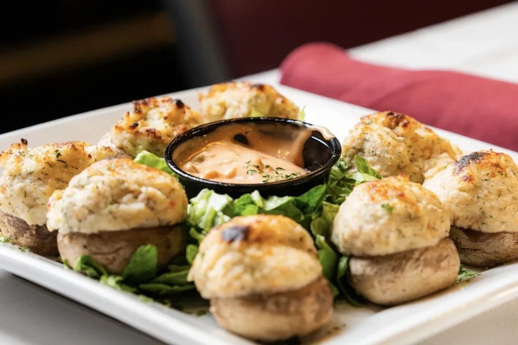 Eight stuffed mushrooms on a white plate with dipping sauce and lettuce.