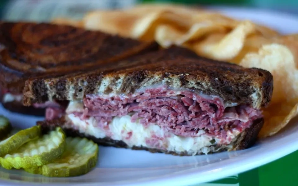 Close-up of a meat sandwich on rye with chips and pickles.