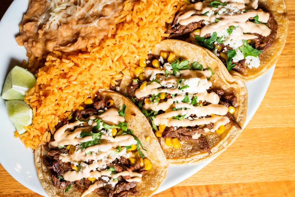 Plate of three carne asada tacos, Spanish rice, refried beans, and lime.