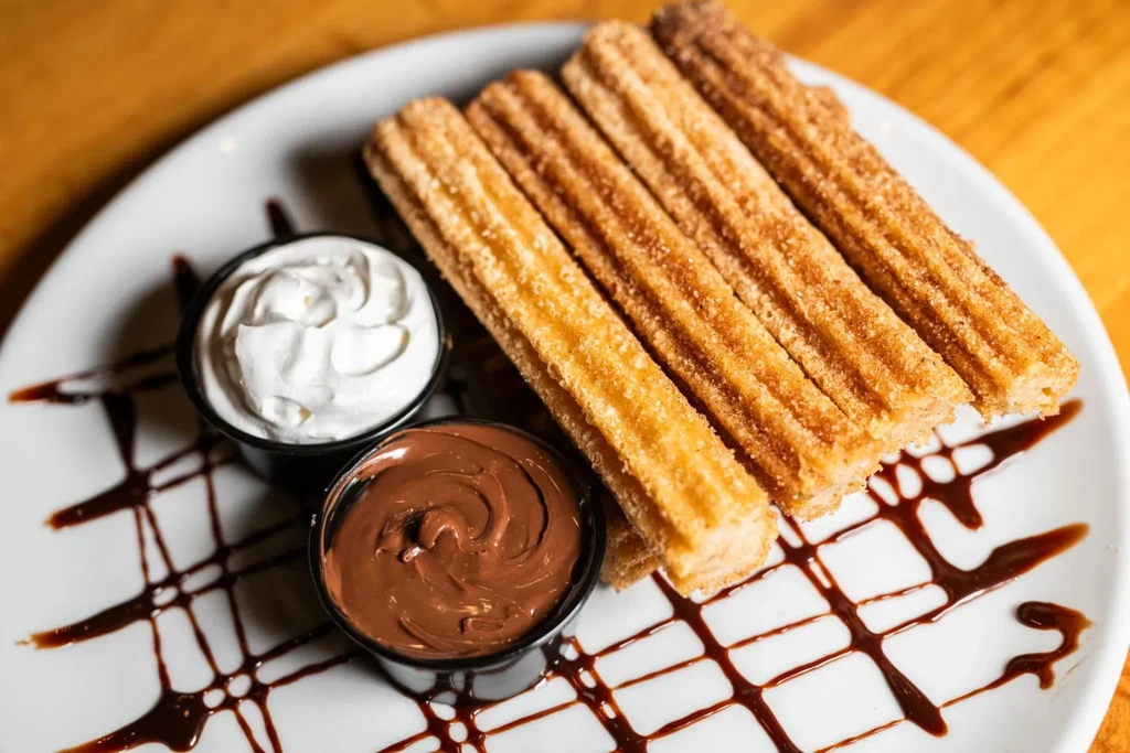 Four golden-brown churros with whipped cream and chocolate dipping sauce.