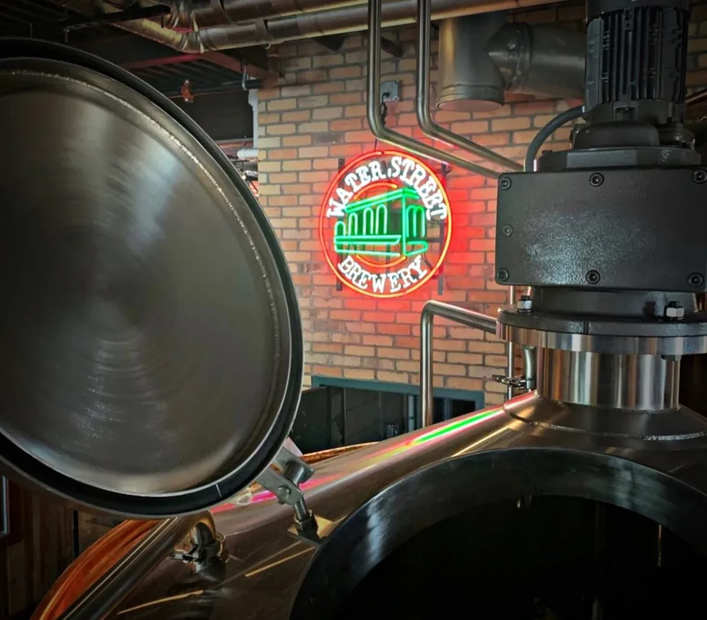 Shiny stainless steel brewing equipment in front of a Water Street Brewery neon sign.