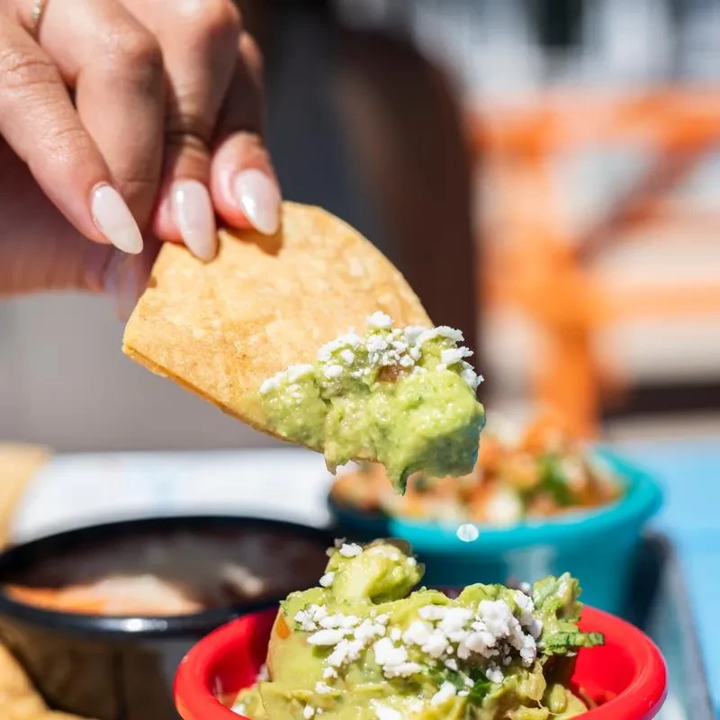 Close-up: hand holding chip in chunky guacamole with crumbled cheese.