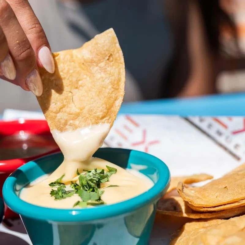 Hand dipping crispy tortilla chip into teal bowl of queso with cilantro.