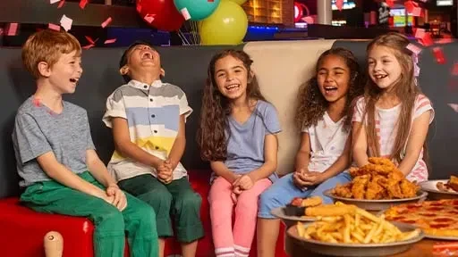 Five laughing children at a table with pizza, chicken, fries, confetti.