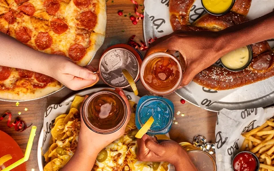 Overhead shot: Diverse hands share pizza, nachos, pretzel, fries, and drinks.