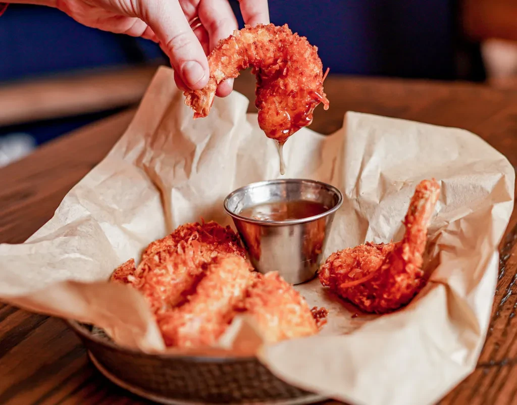 Hand dipping crispy coconut shrimp from basket into amber sauce.