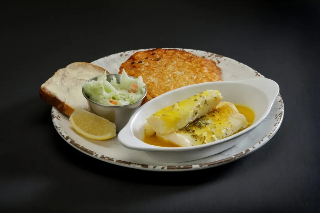 A plate of fish fillets, hash brown, toast, coleslaw, and lemon wedge.