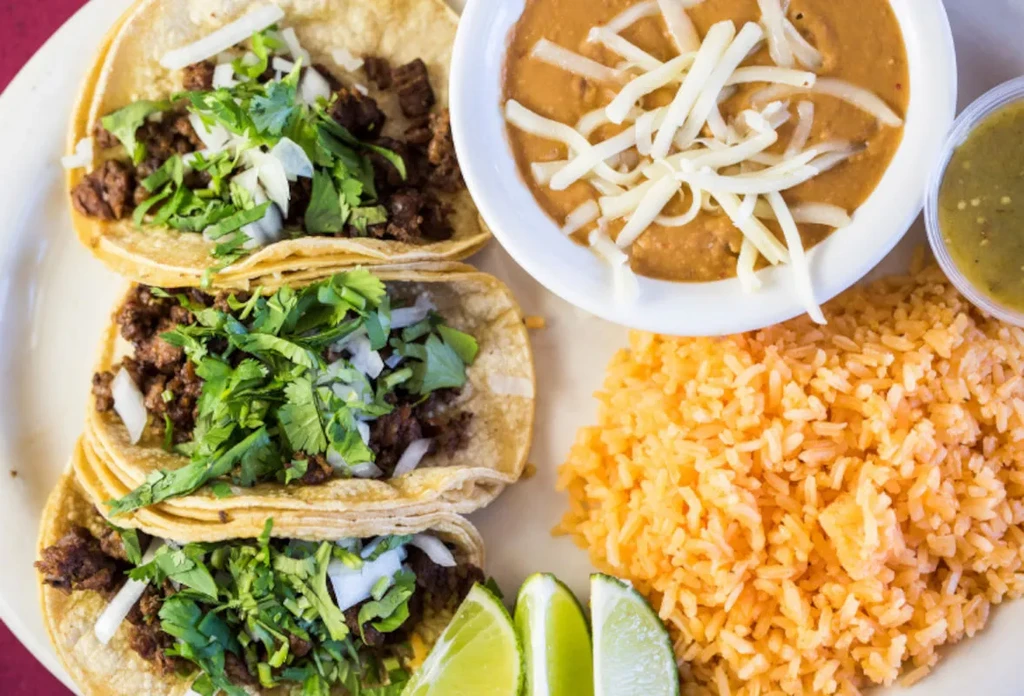 Plate of three street tacos, rice, refried beans, salsa, and limes.