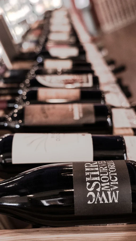 Horizontal wine bottles on a wooden rack, one labeled SHIRAZ MOURVEDRE.