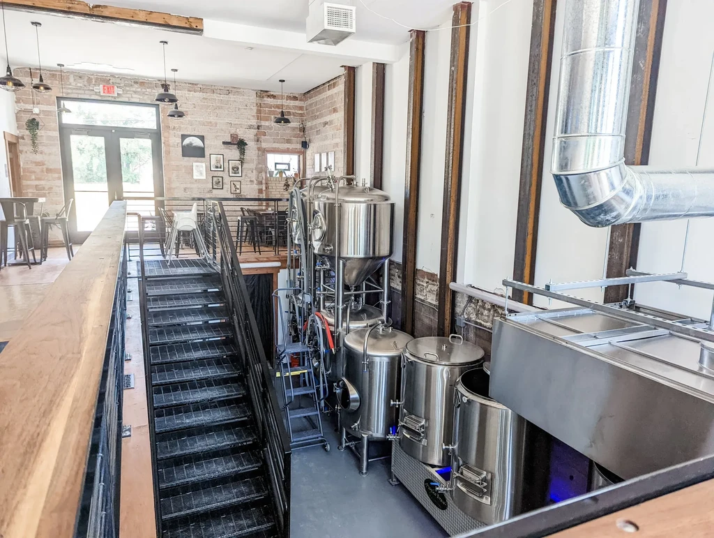 Industrial-chic brewery interior with brewing tanks, staircase, and seating areas.