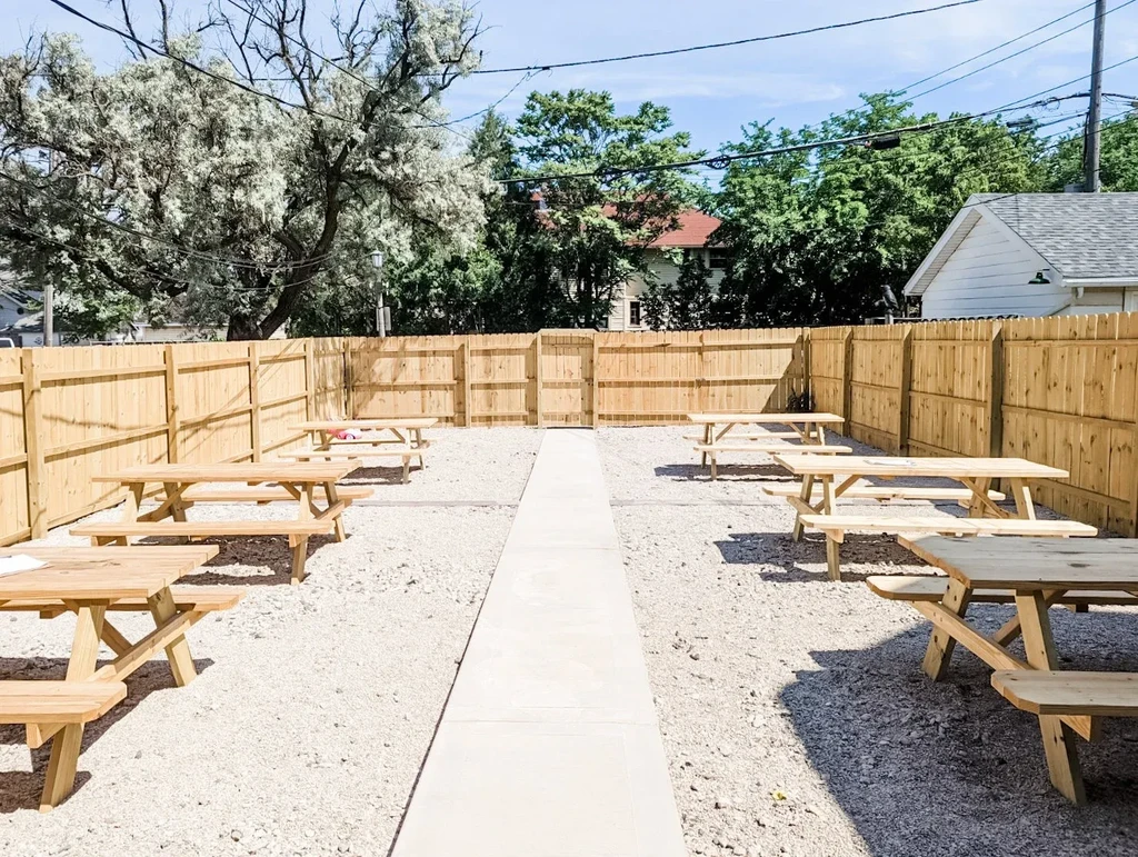 Sunny, fenced outdoor patio with wooden picnic tables on gravel.
