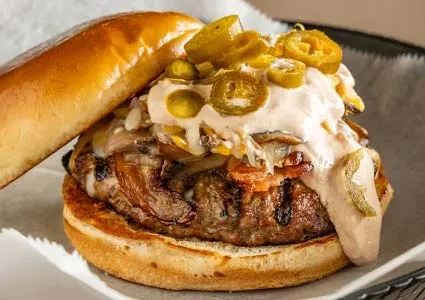 Gourmet burger: thick patty, bacon, cheese, jalapeños, and pinkish-orange sauce.