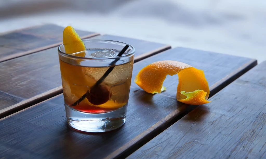 Amber cocktail with cherry, ice, and orange peel on rustic wood.