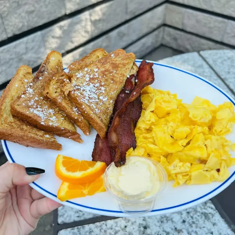 Hearty outdoor breakfast plate with French toast, bacon, eggs, and oranges.