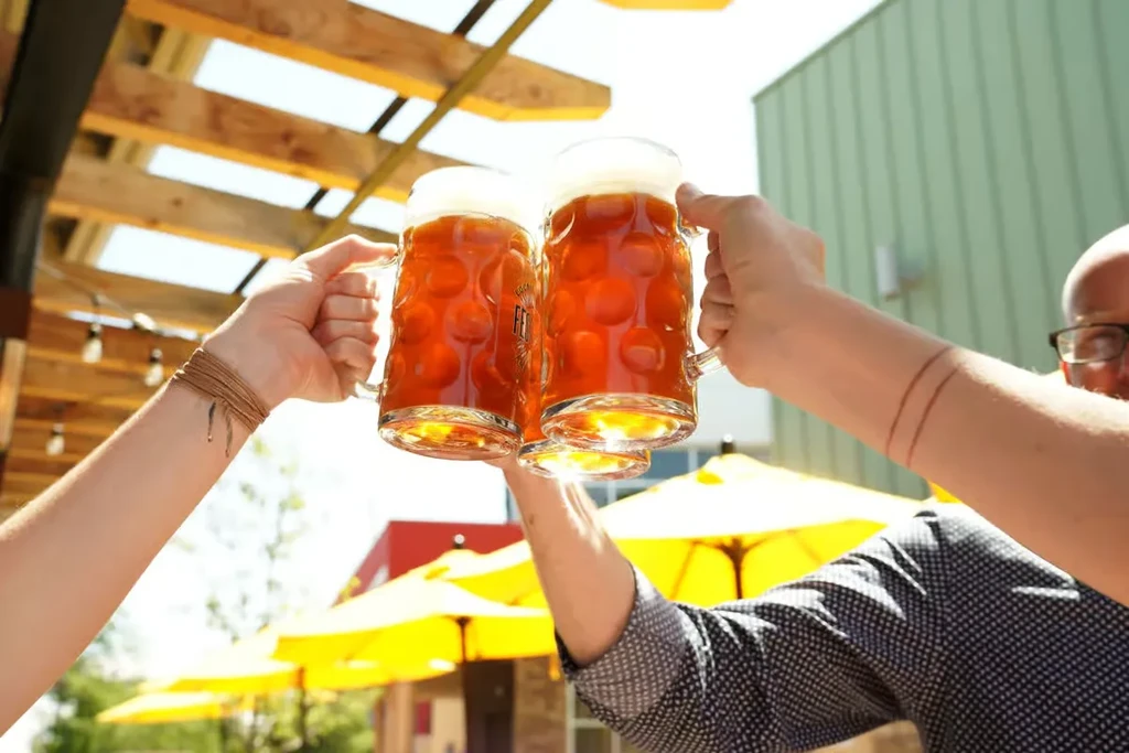 Hands toast two foamy amber beers under a sunny outdoor pergola.