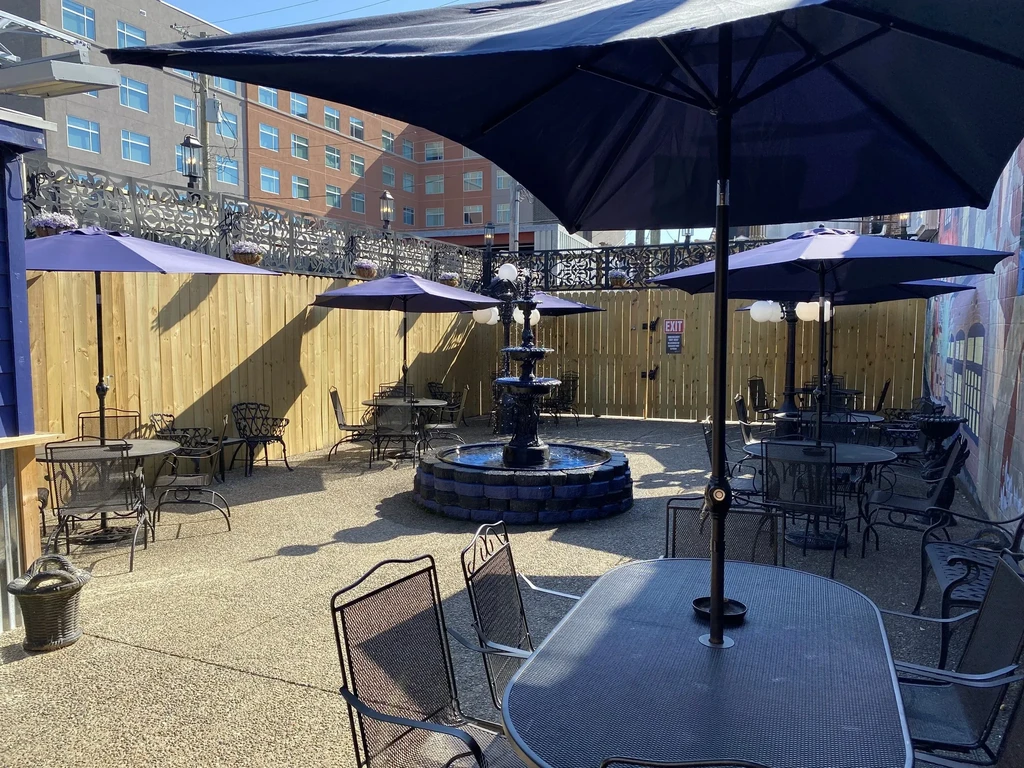 Outdoor restaurant patio with tiered fountain, blue umbrellas, and colorful mural.