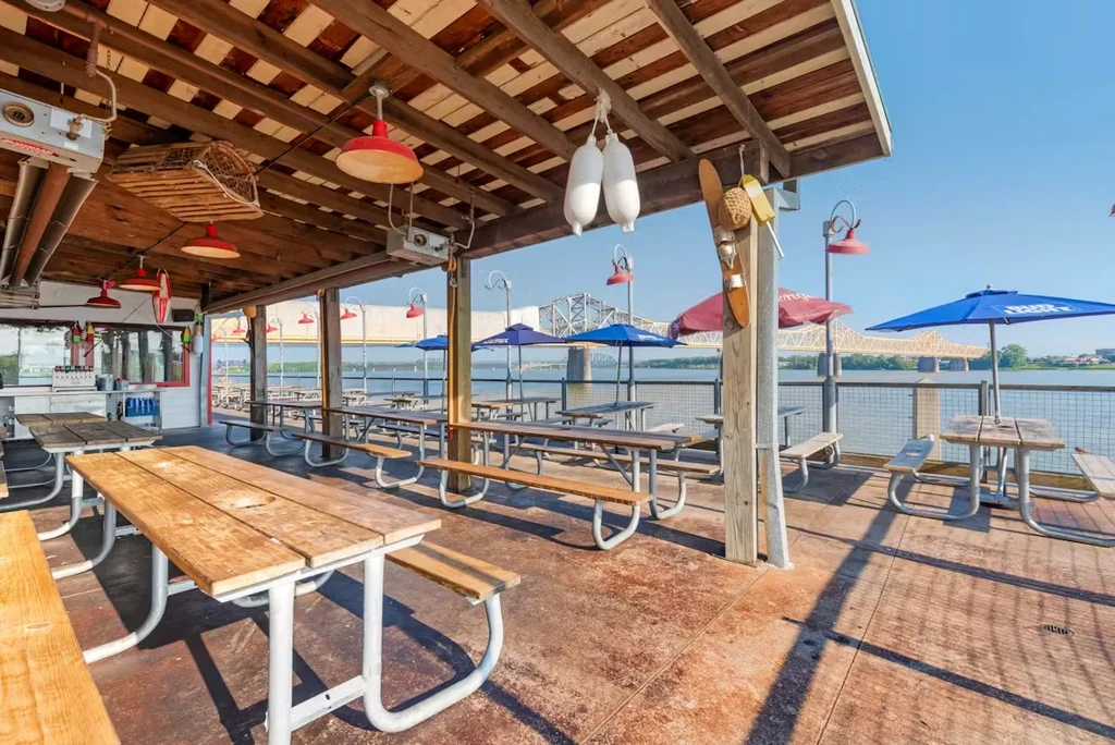 Rustic waterside patio with empty tables, umbrellas, and a large bridge.