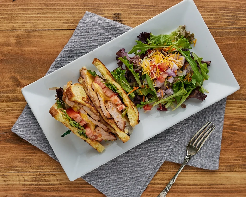 Overhead view: Grilled club sandwich, side salad, fork on wood.