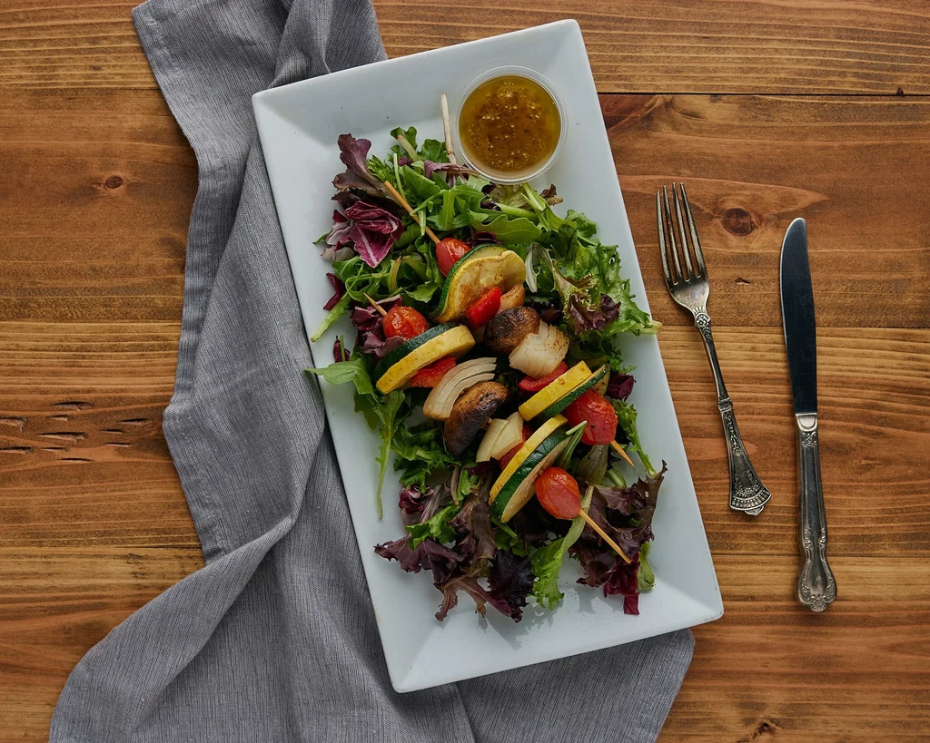 Vibrant salad with grilled vegetable skewers, dressing, and cutlery.