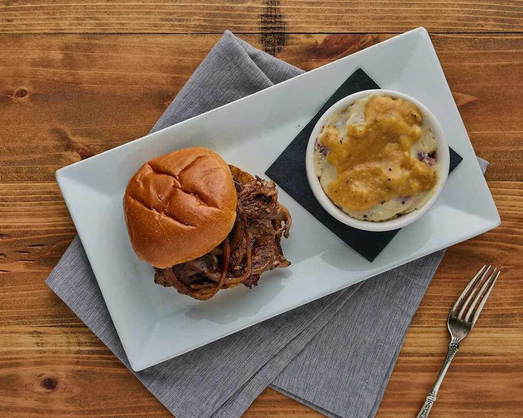 Pulled meat sandwich, mashed potatoes with gravy, and fork on wooden table.