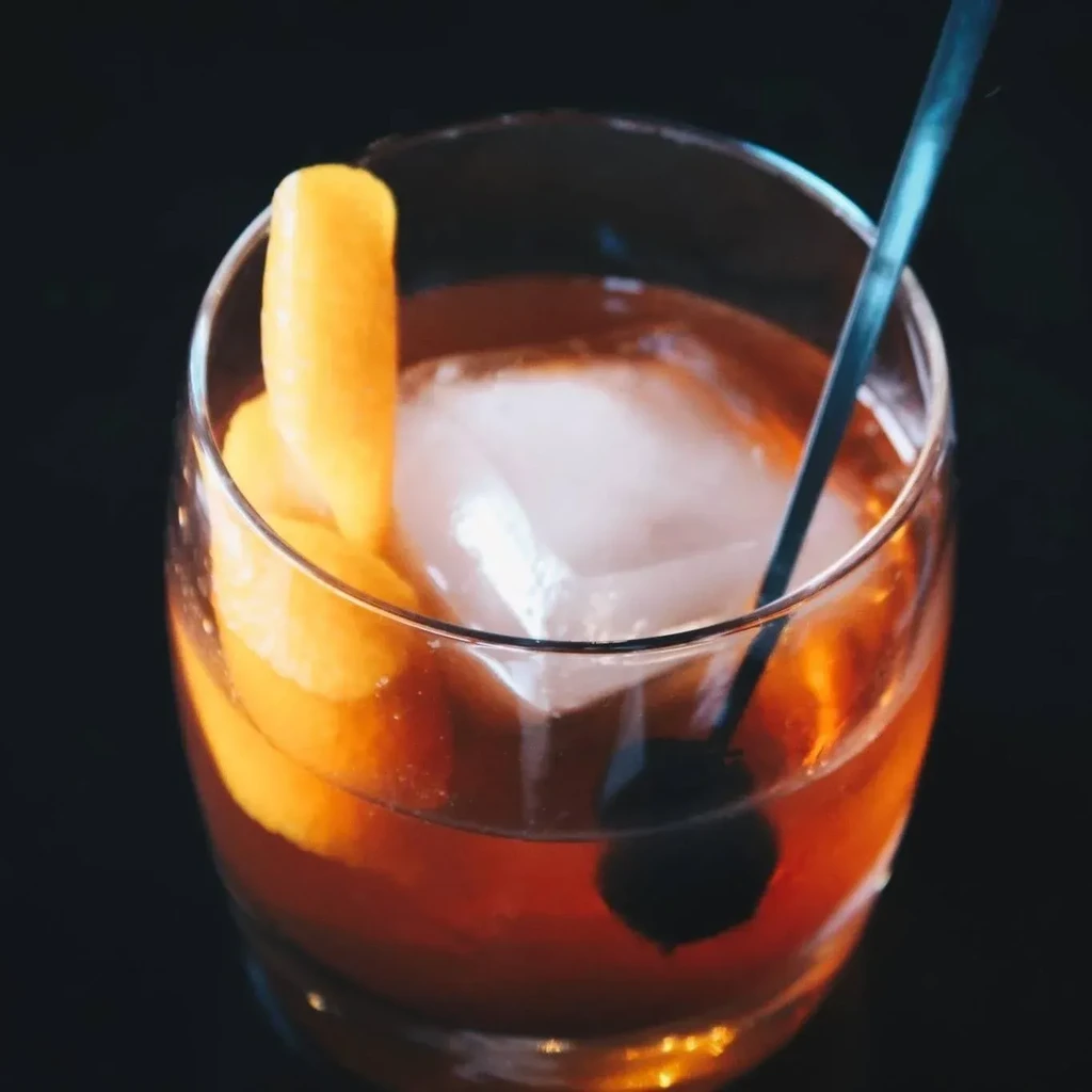 Close-up of amber cocktail with large ice, orange peel, and dark cherry.