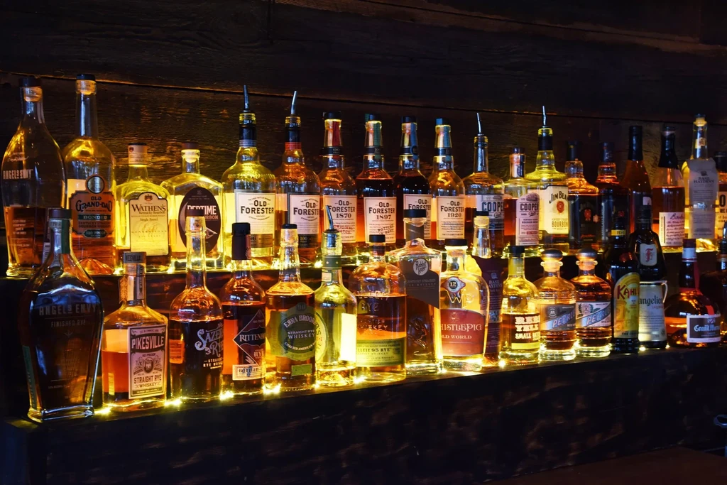 Various whiskey and bourbon bottles glowing on a rustic wooden bar shelf.