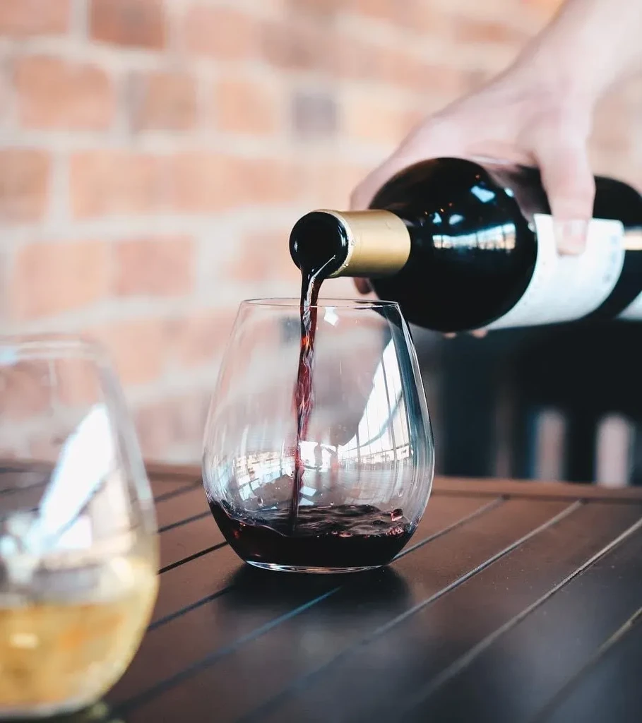 A hand pours dark red wine into a clear stemless glass.